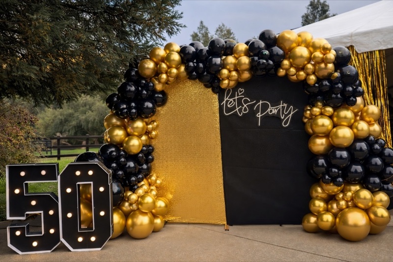 Elegant 50th birthday party setup with black and gold balloon decor and 'Let's Party' sign